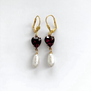 May include: A pair of gold-tone leverback earrings featuring a faceted dark red heart-shaped stone suspended above a white teardrop-shaped pearl. The earrings are symmetrical, with each piece showcasing a deep crimson heart and a smooth white drop.