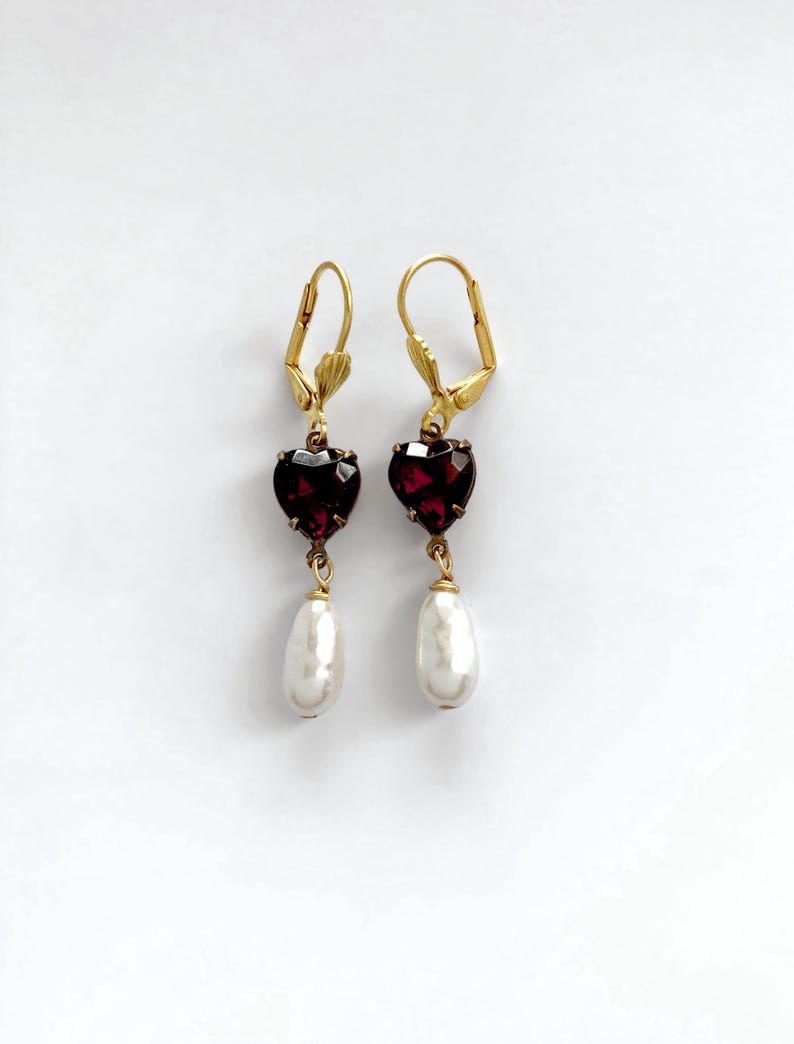 May include: A pair of gold-tone leverback earrings featuring a faceted dark red heart-shaped stone suspended above a white teardrop-shaped pearl. The earrings are symmetrical, with each piece showcasing a deep crimson heart and a smooth white drop.