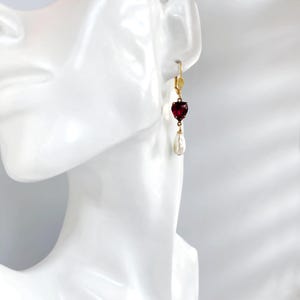 May include: A gold leverback earring featuring a faceted, heart-shaped deep red gemstone suspended above a white, teardrop-shaped pearl. The earring is displayed on a white mannequin ear, showcasing the gold metal setting and the contrast between the red stone and the white pearl.