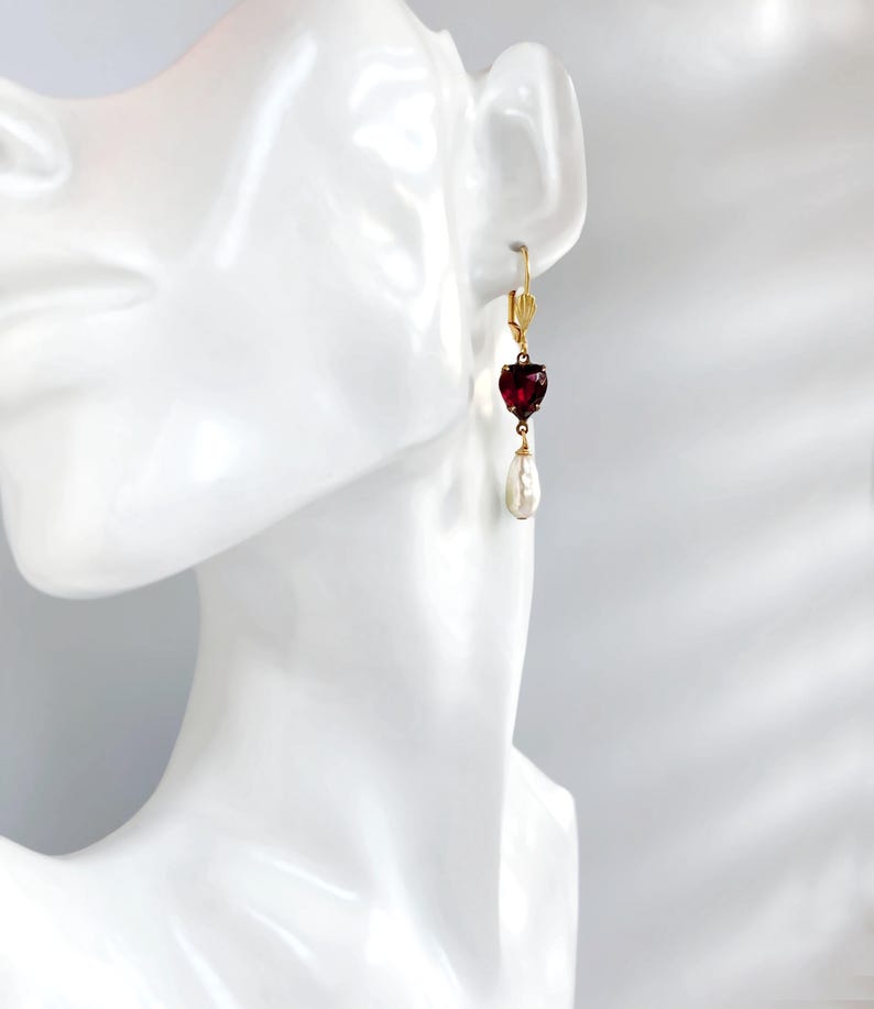 May include: A gold leverback earring featuring a faceted, heart-shaped deep red gemstone suspended above a white, teardrop-shaped pearl. The earring is displayed on a white mannequin ear, showcasing the gold metal setting and the contrast between the red stone and the white pearl.