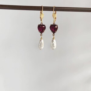 May include: A pair of drop earrings featuring gold-tone leverback closures, each suspending a faceted deep red heart-shaped stone. Below each heart hangs a single white teardrop-shaped pearl, creating a balanced and symmetrical jewellery design.