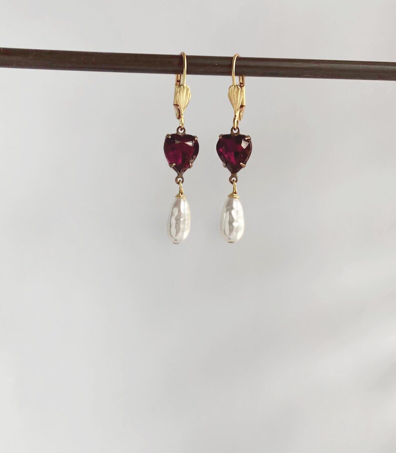 May include: A pair of drop earrings featuring gold-tone leverback closures, each suspending a faceted deep red heart-shaped stone. Below each heart hangs a single white teardrop-shaped pearl, creating a balanced and symmetrical jewellery design.
