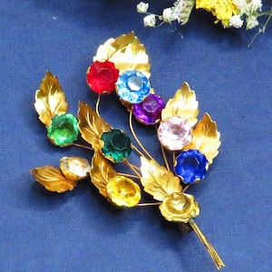 May include: A gold-toned brooch in the shape of a flower bouquet with colorful rhinestones. The brooch features green, blue, red, pink, and yellow rhinestones.