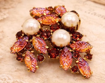 May include: A vintage brooch featuring three large faux pearls and numerous smaller red and orange-toned rhinestones. The brooch has a gold-tone metal setting with leaf-shaped accents. The brooch is set against a backdrop of lace and aged paper.