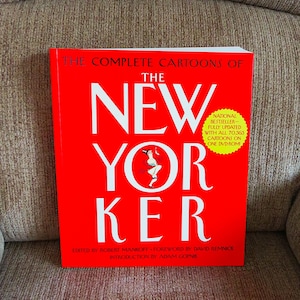 May include: A red book titled "The Complete Cartoons of The New Yorker" with white lettering. The cover features the magazine's logo and a cartoon illustration. A yellow sticker indicates it's a national bestseller with cartoons on DVD-ROM.