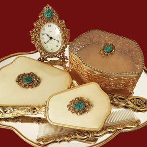 May include: A vintage vanity set on a silver tray. The set includes a small clock, a jewelry box, a hand mirror, a brush, and a comb. Each piece has a gold-tone frame and a cream-colored surface with a decorative green stone.