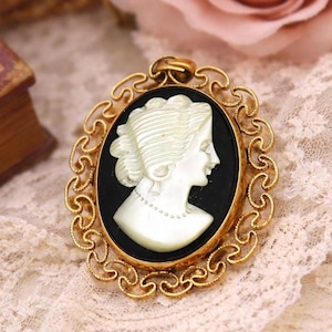 May include: Oval cameo pendant with a profile of a woman in white against a black background, set in a gold-toned filigree frame. The pendant is likely vintage or antique, showcasing a classic jewelry design.