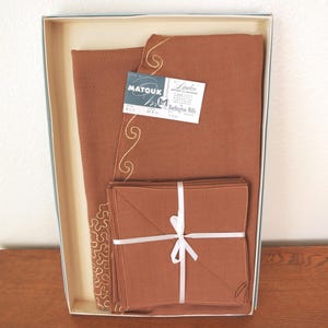 May include: A set of brown linens, including a folded tablecloth and napkins, presented in a box. The tablecloth features gold embroidered detailing. The napkins are tied with a white ribbon. The box contains tags that read "Matouk" and "Linden".