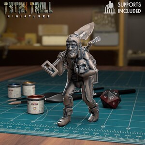 Könnte beinhalten: Eine graue Miniaturfigur eines alten Mannes mit Schaufel und Schädel, mit dem Text "TITAN TROLL MINIATURES". Die Figur steht auf einer grünen Oberfläche mit Malutensilien und einem Würfel. Der Text "SUPPORTS INCLUDED" ist ebenfalls sichtbar.