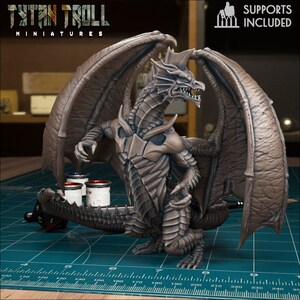 Könnte beinhalten: Eine detaillierte 3D-gedruckte Miniatur eines grauen Drachen mit ausgebreiteten Flügeln. Der Drache ist in einer stehenden Position mit dem Kopf zur Seite gedreht. Die Miniatur steht auf einer grünen Schneidematte mit einem Lineal und Farbtöpfen.