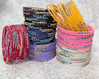 May include: A collection of colorful bangles in shades of red, black, navy, purple, yellow, pink, and white. Each bangle is adorned with decorative accents of gold, blue, and other colors. The bangles are stacked in piles, showcasing their vibrant designs.