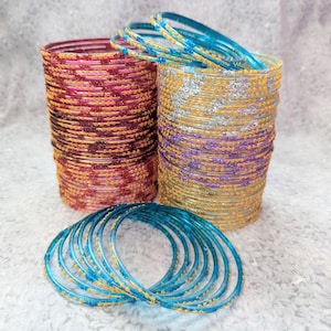 May include: Stacks of colorful bangles in shades of red, purple, yellow, and blue. The bangles have a metallic gold accent. Some bangles are stacked in cylindrical shapes, while others are scattered in the foreground.