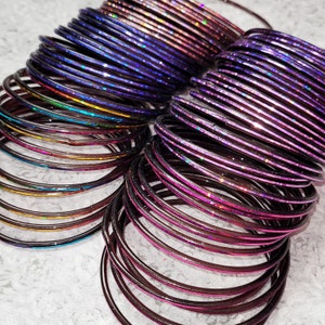 May include: A collection of colorful bangles in shades of purple, pink, and iridescent hues. The bangles are stacked together, creating a layered effect. The light reflects off the shiny surfaces, highlighting the various colors and textures.