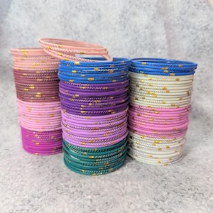 Puede incluir: Montones de pulseras coloridas en tonos rosa, morado, azul, verde y blanco. Cada pulsera está adornada con detalles dorados. Las pulseras están apiladas en montones ordenados, mostrando una variedad de colores y diseños.