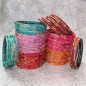 May include: A selection of colourful bangles in stacks. The bangles are in shades of teal, pink, purple, red, and orange, with gold floral patterns. Some bangles are stacked, while others are in a pile. The bangles are made of glass.