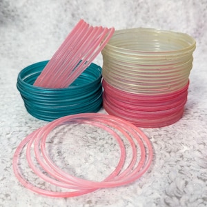 Puede incluir: Una colección de pulseras coloridas en tonos rosa, turquesa y transparente. Las pulseras están apiladas en grupos, algunas dispuestas en forma circular. La imagen muestra la variedad de colores y estilos.