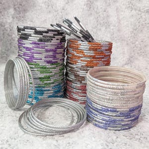 May include: A collection of colorful bangles in various stacks and arrangements. The bangles feature silver, black, purple, green, orange, and red accents. Some bangles are stacked, while others are arranged in loose piles. The bangles are made of a shiny material.