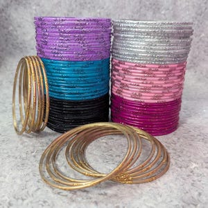 May include: Stacks of colorful bangles in shades of purple, teal, black, silver, pink, and magenta. Several gold-colored bangles are arranged in front of the stacks. The bangles have a shiny, metallic finish with glitter accents, perfect for adding a touch of sparkle.
