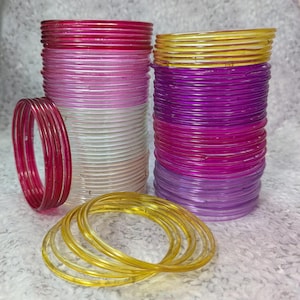 Może przedstawiać: Kolekcja kolorowych bransoletek w odcieniach różu, fioletu i żółci. Bransoletki ułożone są w schludne stosy, niektóre ułożone w okrąg. Przezroczyste bransoletki tworzą żywą i przyciągającą wzrok ekspozycję.