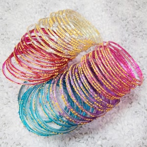 Pode incluir: Uma coleção de pulseiras coloridas em tons de ouro, vermelho, rosa, roxo e azul-turquesa. As pulseiras são adornadas com detalhes decorativos, criando uma exibição festiva e atraente. As pulseiras estão empilhadas juntas.