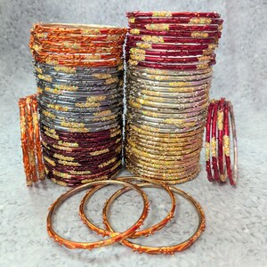 May include: Stacks of colorful bangles in shades of orange, silver, gold, and red. Some bangles have decorative embellishments. Several loose bangles are in the foreground. The bangles are likely for fashion or cultural wear.