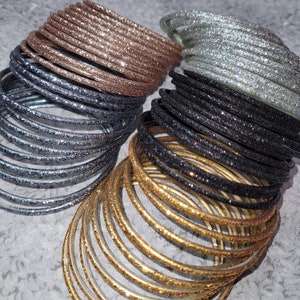 Op de afbeelding: Een verzameling glitterarmbanden in verschillende kleuren, waaronder zilver, zwart, goud en roségoud. De armbanden zijn in meerdere stapels gerangschikt, wat hun glinsterende textuur en ontwerp benadrukt. Deze armbanden zijn perfect om een vleugje sprankeling aan elke outfit toe te voegen.