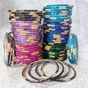 May include: A collection of colourful bangles in black, purple, pink, teal, and green. Each bangle features gold glitter accents. The bangles are arranged in stacks and individually, highlighting the range of colours and designs available.