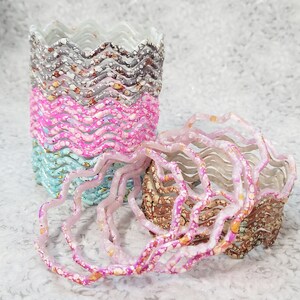 May include: A stack of wavy-edged bangles in various colors, including pink, blue, and brown, with a speckled pattern. Several bangles are arranged in a stack, while others are scattered nearby. The bangles have a unique, textured design.