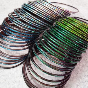 May include: A collection of thin, iridescent bangles in shades of blue, green, and silver. The bangles have a subtle sparkle and are stacked in multiple piles, creating a layered effect. The bangles are a fashion accessory.