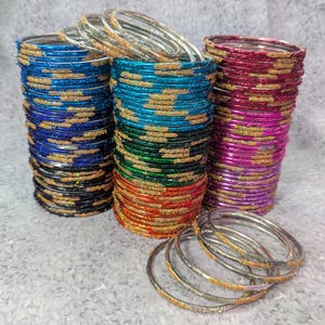 May include: Stacks of colorful bangles in shades of blue, teal, green, orange, and pink. Each bangle is adorned with a gold glitter design. A few loose bangles are in the foreground. The bangles are a traditional jewelry item.