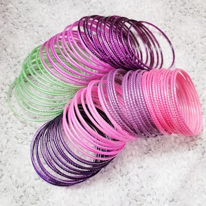Puede incluir: Una colección de pulseras coloridas en tonos rosa, morado y verde. Las pulseras están dispuestas en pilas curvas, mostrando su diseño liso y redondeado. La luz se refleja en las superficies, creando una vibrante exhibición de accesorios.