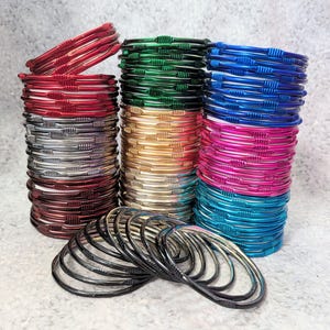 May include: Stacks of colorful bangles in red, silver, gold, green, blue, pink, and teal. The bangles are thin, with a textured design. A few bangles are arranged separately in the foreground. The bangles are stacked in multiple piles.