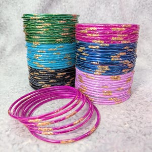 May include: Stacks of colorful bangles in green, turquoise, black, blue, and pink, with gold glitter accents. A few pink bangles are scattered in front of the stacks. The bangles are on a white, textured surface.