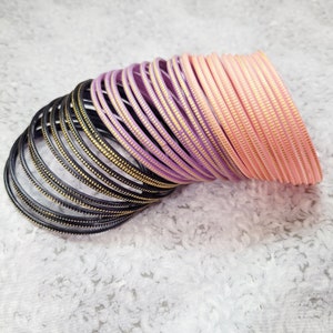 Op de afbeelding: Een verzameling armbanden in verschillende kleuren, waaronder zwart, lavendel en perzik. Elke armband heeft een getextureerd oppervlak met goudkleurige accenten. De armbanden zijn op elkaar gestapeld, wat een visueel aantrekkelijke presentatie creëert.