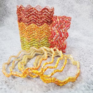 Peut inclure: Ensemble de récipients en résine aux bords ondulés et colorés, dans les tons de rouge, orange et jaune. Plusieurs bagues en résine assorties à bords ondulés sont devant les récipients. La résine présente un motif tacheté.