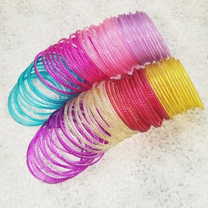 Puede incluir: Una colección de pulseras coloridas en tonos turquesa, rosa, morado, rojo y amarillo. Las pulseras están apiladas en múltiples filas, creando una exhibición vibrante. Las pulseras transparentes tienen una superficie texturizada y están dispuestas sobre un fondo blanco texturizado.