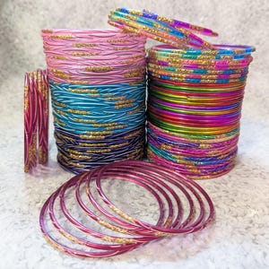 May include: Stacks of colorful bangles in pink, teal, and purple with gold glitter accents. Additional bangles are arranged in stacks and scattered on the surface. The bangles are made of glass and are a traditional Indian accessory.