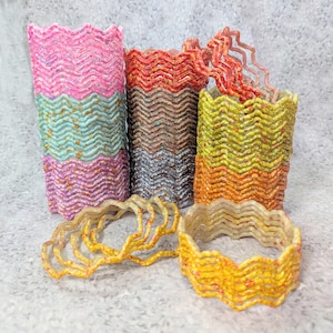 Op de afbeelding: Een verzameling kleurrijke, golvende decoratieve containers. De containers zijn gestapeld en hebben een verscheidenheid aan kleuren, waaronder roze, blauw, oranje, geel en bruin. Sommige containers hebben gouden vlekken. Er zijn ook verschillende losse, golvende ringen.