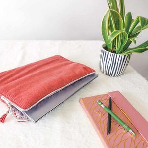 Könnte beinhalten: Eine korallenfarbene Laptop-Hülle aus Cord mit einem Quasten-Reißverschluss, die einen silbernen Laptop enthält. Ein rosa Notizbuch mit goldenem Blättermuster und zwei Stiften liegen neben der Hülle. Eine Topf-Schlangenpflanze befindet sich im Hintergrund.