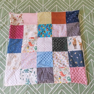 May include: A patchwork quilt with a variety of patterned squares. The squares feature prints of deer, dinosaurs, polka dots, and floral designs. Colours include pink, blue, brown, and yellow. The quilt is likely for a baby or child.
