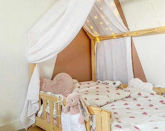 May include: A wooden toddler bed with a canopy, adorned with fairy lights. The bed features a white and pink heart-patterned duvet cover and pillow. A plush bunny and heart-shaped pillow add to the cozy aesthetic. The canopy is draped with white fabric.
