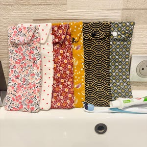 Peut inclure: Six étuis à brosse à dents en tissu aux motifs et couleurs variés. Les étuis présentent des motifs floraux, en forme de cœur et géométriques en rose, rouge, blanc, jaune et noir. Une brosse à dents et du dentifrice sont visibles sur l'évier.