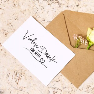 K&ouml;nnte beinhalten: Eine wei&szlig;e Karte mit dem Text "Vielen Dank f&uuml;r alles" in schwarzer Schrift mit einem kleinen Herzsymbol. Die Karte befindet sich in einem braunen Umschlag mit einer wei&szlig;en Blume und kleinen rosa Bl&uuml;ten, die herausragen.