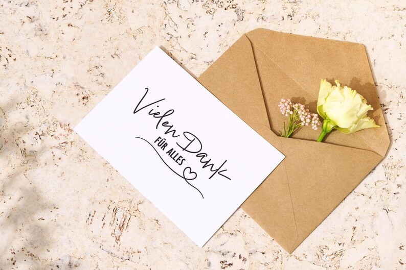 K&ouml;nnte beinhalten: Eine wei&szlig;e Karte mit dem Text "Vielen Dank f&uuml;r alles" in schwarzer Schrift mit einem kleinen Herzsymbol. Die Karte befindet sich in einem braunen Umschlag mit einer wei&szlig;en Blume und kleinen rosa Bl&uuml;ten, die herausragen.