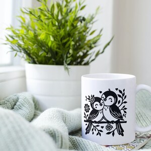 May include: White ceramic mug with a black silhouette design of two birds perched on a branch with flowers and hearts. The mug is sitting on a green knitted blanket.