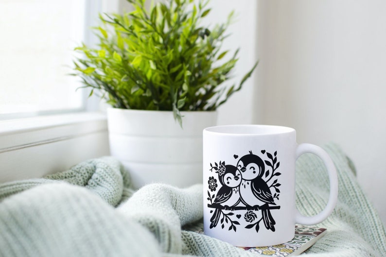 May include: White ceramic mug with a black silhouette design of two birds perched on a branch with flowers and hearts. The mug is sitting on a green knitted blanket.