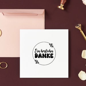K&ouml;nnte beinhalten: Eine wei&szlig;e Karte mit einem schwarzen Kreis und dem Text "Ein herzliches DANKE" im Kreis. Die Karte ist von einem rosa Umschlag und einem goldenen Siegelwachs umgeben.