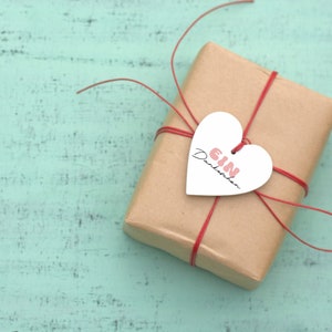 K&ouml;nnte beinhalten: Eine braune Geschenkbox aus Papier, die mit roter Schnur gebunden ist und einen wei&szlig;en herzf&ouml;rmigen Anh&auml;nger mit der Aufschrift "EIN Dankesch&ouml;n" in rosa Schrift.