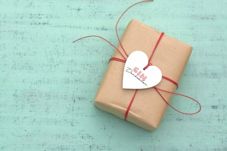 K&ouml;nnte beinhalten: Eine braune Geschenkbox aus Papier, die mit roter Schnur gebunden ist und einen wei&szlig;en herzf&ouml;rmigen Anh&auml;nger mit der Aufschrift "EIN Dankesch&ouml;n" in rosa Schrift.