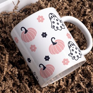 Puede incluir: Taza de cer&aacute;mica blanca con tem&aacute;tica de Halloween, con calabazas rosas, flores negras e ilustraciones de fantasmas. La taza se presenta en una caja de regalo blanca con relleno de papel marr&oacute;n.